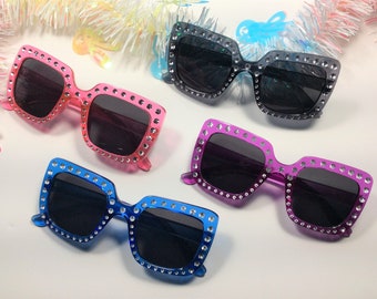 Puede incluir: Cuatro pares de gafas de sol cuadradas con pedrería alrededor de las monturas. Las gafas de sol son de color rosa, morado, azul y negro.