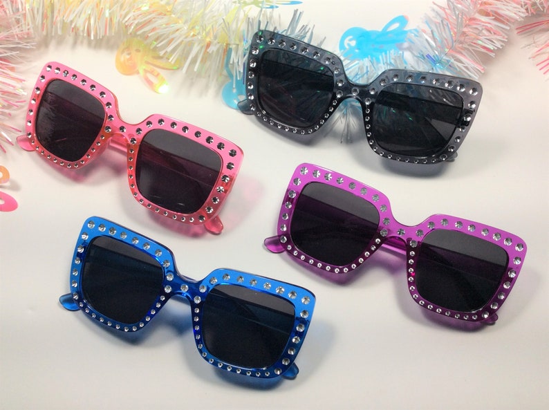 May include: Four pairs of square-shaped sunglasses with rhinestones around the frames. The sunglasses are in pink, purple, blue, and black.