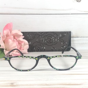 May include: Black half-frame reading glasses with green and yellow detailing and decorative rhinestones. A black case with a rose pattern is behind the glasses. A pink flower is in the background.