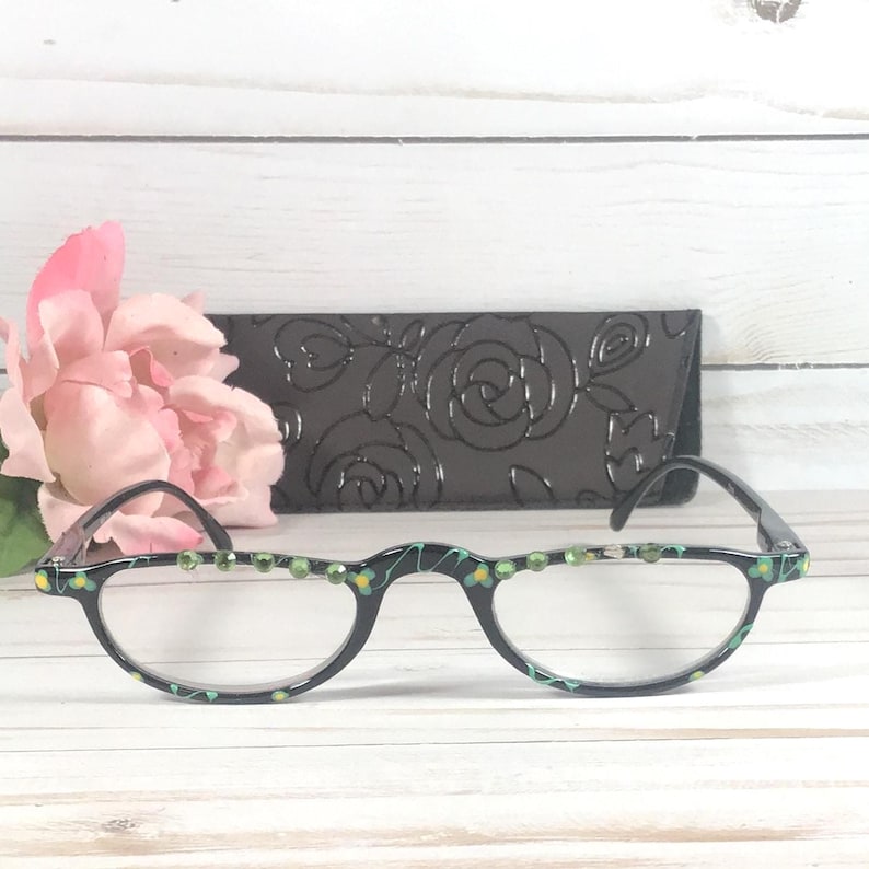 May include: Black half-frame reading glasses with green and yellow detailing and decorative rhinestones. A black case with a rose pattern is behind the glasses. A pink flower is in the background.