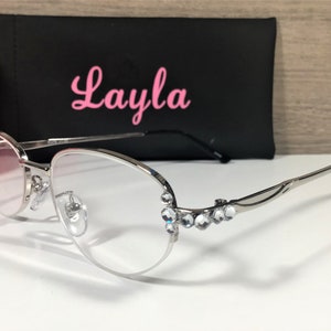 K&ouml;nnte beinhalten: Brille mit silbernem Rahmen und klaren Gl&auml;sern. Die Fassungen weisen eine dekorative Verzierung aus klaren Strasssteinen auf. Im Hintergrund befindet sich ein schwarzes Etui mit dem Namen "Layla" in rosa Schrift.