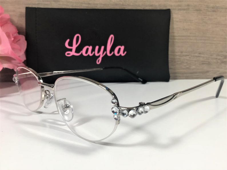 K&ouml;nnte beinhalten: Brille mit silbernem Rahmen und klaren Gl&auml;sern. Die Fassungen weisen eine dekorative Verzierung aus klaren Strasssteinen auf. Im Hintergrund befindet sich ein schwarzes Etui mit dem Namen "Layla" in rosa Schrift.