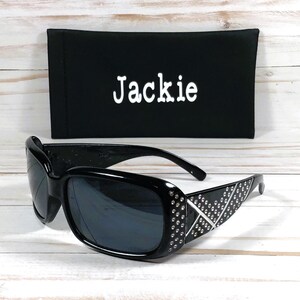 K&ouml;nnte beinhalten: Schwarze Sonnenbrille mit Strasssteinen auf dem Gestell und einem schwarzen Stoffetui mit dem Namen "Jackie" bestickt.