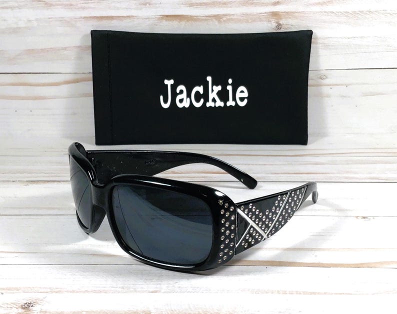 K&ouml;nnte beinhalten: Schwarze Sonnenbrille mit Strasssteinen auf dem Gestell und einem schwarzen Stoffetui mit dem Namen "Jackie" bestickt.