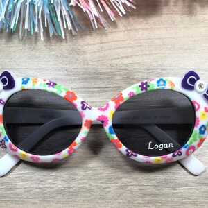 May include: White oval sunglasses with a floral print and purple bows on the sides. The sunglasses have dark lenses and the name "Logan" is printed on the right lens.