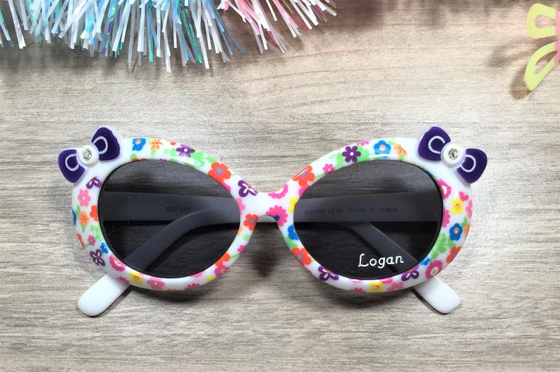 May include: White oval sunglasses with a floral print and purple bows on the sides. The sunglasses have dark lenses and the name "Logan" is printed on the right lens.