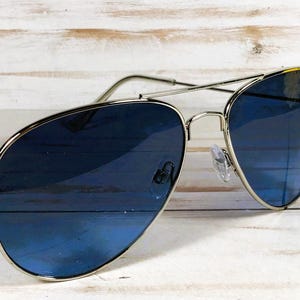 May include: Aviator sunglasses featuring a silver frame and gradient blue lenses. The lenses transition from a darker blue at the top to a lighter shade at the bottom. The sunglasses are set against a light wood grain background.