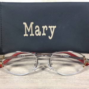 Puede incluir: Gafas con montura plateada, detalles rojos y estuche negro. El estuche lleva el nombre "Mary". Las gafas tienen un dise&ntilde;o de media montura. Est&aacute;n sobre una superficie de madera.