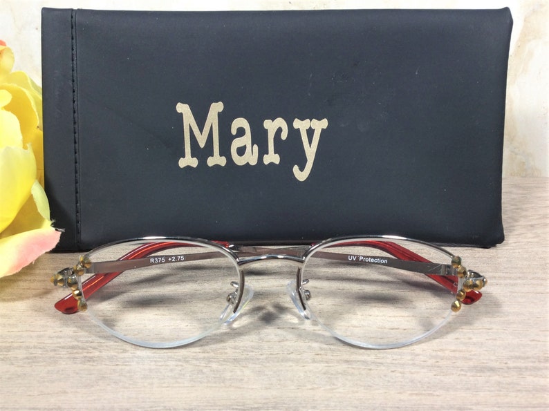 Puede incluir: Gafas con montura plateada, detalles rojos y estuche negro. El estuche lleva el nombre "Mary". Las gafas tienen un dise&ntilde;o de media montura. Est&aacute;n sobre una superficie de madera.