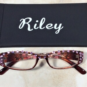 May include: Brown-framed reading glasses with pink rhinestones along the top rim. The lenses have a floral pattern. A black case with the name "Riley" in white script is behind the glasses.