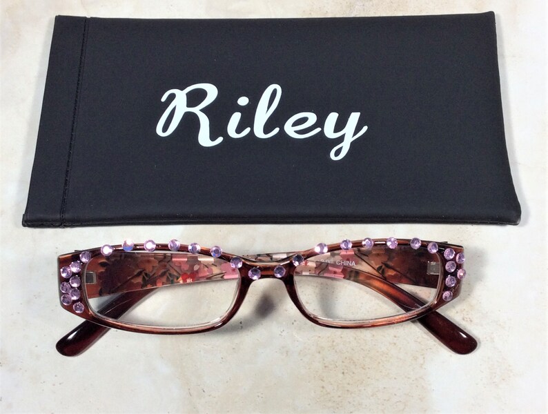 May include: Brown-framed reading glasses with pink rhinestones along the top rim. The lenses have a floral pattern. A black case with the name "Riley" in white script is behind the glasses.