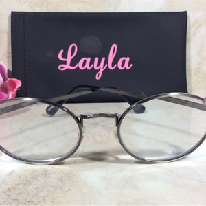 Puede incluir: Gafas de forma ovalada con montura de color plomo y lentes transparentes. La montura est&aacute; adornada con pedrer&iacute;a rosa en las sienes. Detr&aacute;s de las gafas hay un estuche negro con el nombre "Layla" en escritura rosa. Una flor rosa en el fondo.