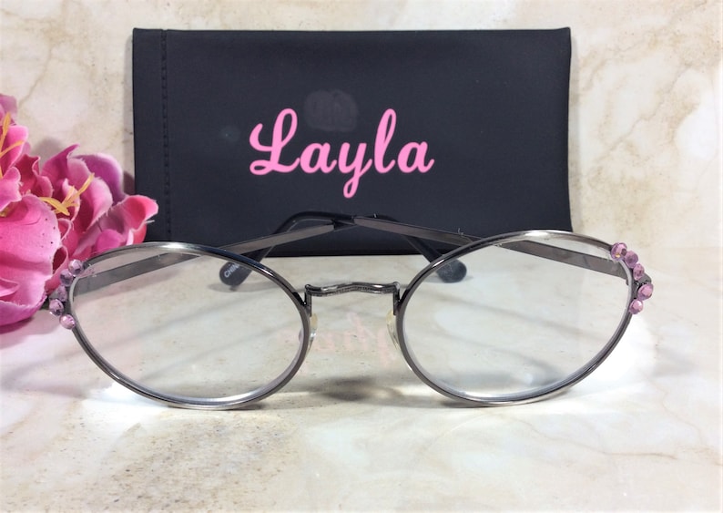 Puede incluir: Gafas de forma ovalada con montura de color plomo y lentes transparentes. La montura est&aacute; adornada con pedrer&iacute;a rosa en las sienes. Detr&aacute;s de las gafas hay un estuche negro con el nombre "Layla" en escritura rosa. Una flor rosa en el fondo.