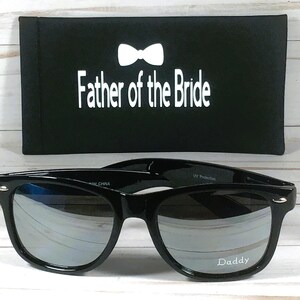 May include: Black sunglasses with mirrored lenses and a black case. The case has a white bow tie and the text "Father of the Bride". The sunglasses have the word "Daddy" printed on the right lens.