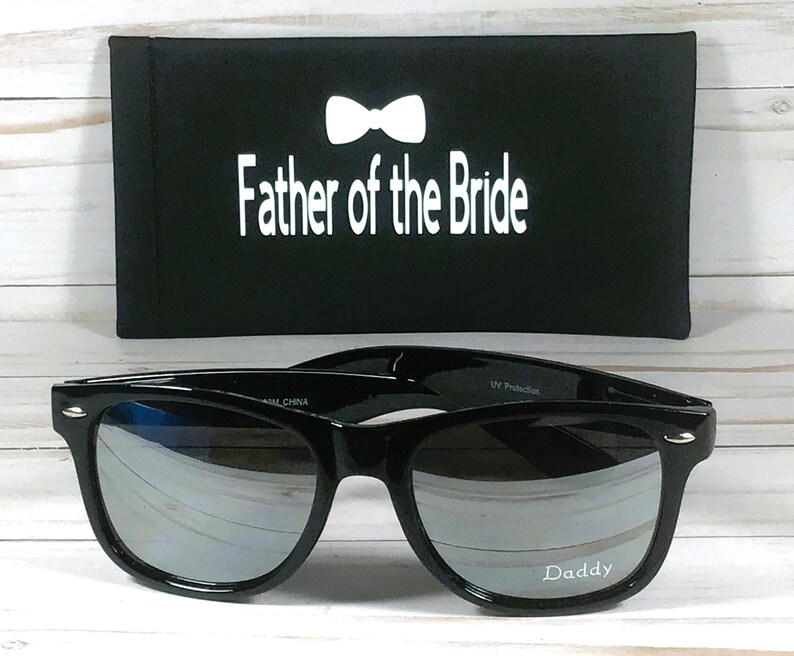 May include: Black sunglasses with mirrored lenses and a black case. The case has a white bow tie and the text "Father of the Bride". The sunglasses have the word "Daddy" printed on the right lens.