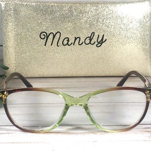 Pu&ograve; includere: Occhiali con montatura sfumata verde e marrone, davanti a un astuccio dorato. L'astuccio &egrave; impresso con un motivo a pois e il nome "Mandy" in corsivo.