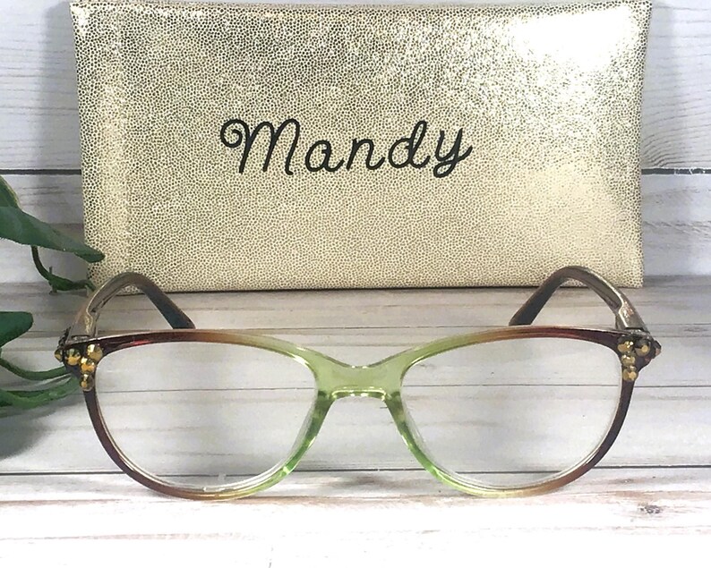 Pu&ograve; includere: Occhiali con montatura sfumata verde e marrone, davanti a un astuccio dorato. L'astuccio &egrave; impresso con un motivo a pois e il nome "Mandy" in corsivo.