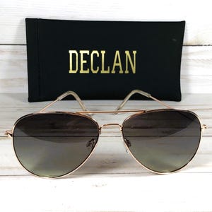May include: Rose gold aviator sunglasses with gradient lenses. The sunglasses are paired with a black case with the name "DECLAN" in gold lettering.