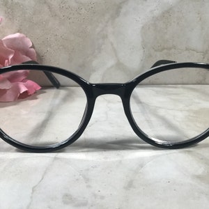 May include: Black round-frame eyeglasses with clear lenses. The frames are adorned with clusters of small, pink rhinestones at the temples. The glasses are set against a soft, blurred background with a pink flower.