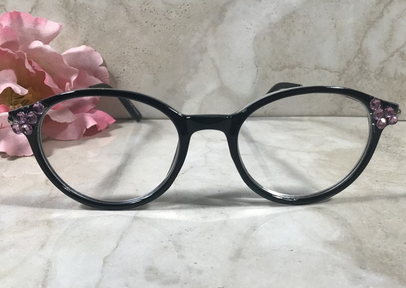 May include: Black round-frame eyeglasses with clear lenses. The frames are adorned with clusters of small, pink rhinestones at the temples. The glasses are set against a soft, blurred background with a pink flower.