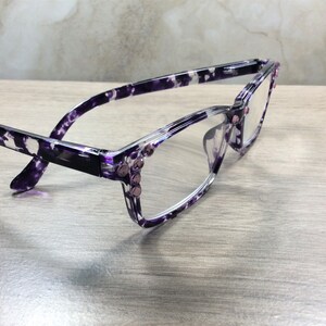 K&ouml;nnte beinhalten: Eine Brille mit einem Rahmen in einem lila-wei&szlig;en Sprenkelmuster. Die Fassung ist mit kleinen, rosa Strasssteinen an den Scharnieren verziert. Die Gl&auml;ser sind klar, und die Brille liegt auf einer hellen Holzmaserung.