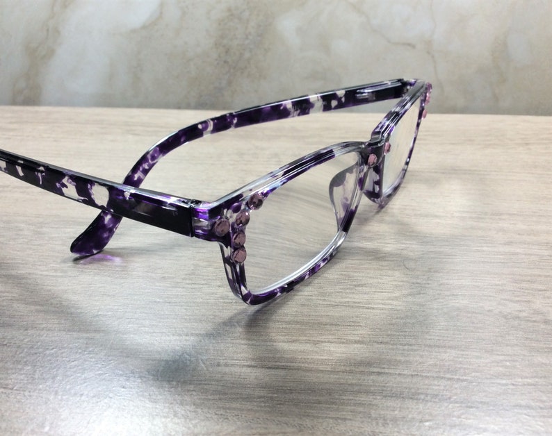K&ouml;nnte beinhalten: Eine Brille mit einem Rahmen in einem lila-wei&szlig;en Sprenkelmuster. Die Fassung ist mit kleinen, rosa Strasssteinen an den Scharnieren verziert. Die Gl&auml;ser sind klar, und die Brille liegt auf einer hellen Holzmaserung.