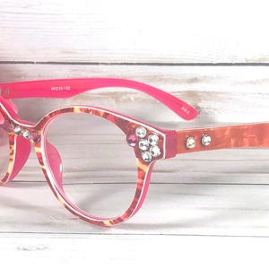 May include: Eyeglasses with a pink and orange marbled pattern frame. The temples are bright pink on the inside and marbled on the outside. The front corners and temples are decorated with clear and pink rhinestones. The frame features 49-18-138 text.