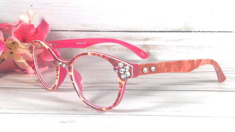 May include: Eyeglasses with a pink and orange marbled pattern frame. The temples are bright pink on the inside and marbled on the outside. The front corners and temples are decorated with clear and pink rhinestones. The frame features 49-18-138 text.