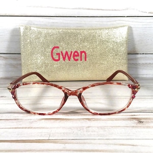 May include: A pair of eyeglasses with a red and white marbled pattern on the frames, featuring small red crystal accents at the hinges. Behind the glasses sits a rectangular gold glitter case with the name Gwen printed in red lettering.