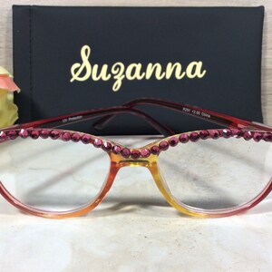 May include: Cat-eye reading glasses with a gradient amber and red frame, embellished with pink rhinestones along the top rim. The glasses are paired with a black case with the name "Suzanna" in gold script.