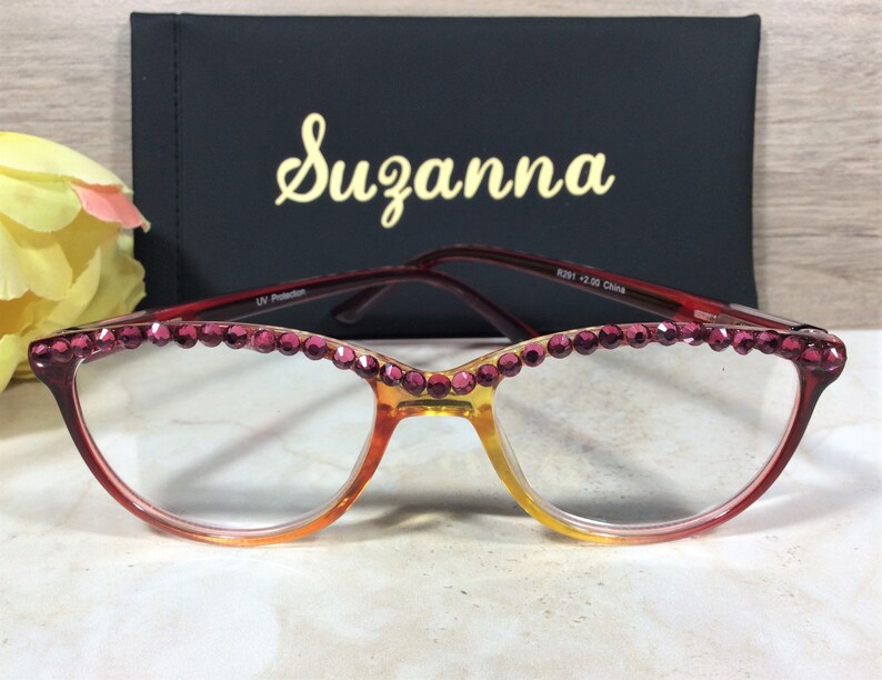 May include: Cat-eye reading glasses with a gradient amber and red frame, embellished with pink rhinestones along the top rim. The glasses are paired with a black case with the name "Suzanna" in gold script.