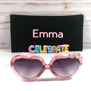 May include: Pink heart-shaped sunglasses with glitter and pink flower accents. The sunglasses come with a black fabric case that has the name "Emma" and the word "CELEBRATE" printed on it.