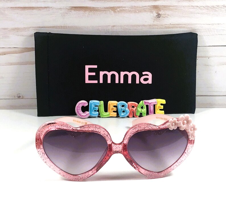May include: Pink heart-shaped sunglasses with glitter and pink flower accents. The sunglasses come with a black fabric case that has the name "Emma" and the word "CELEBRATE" printed on it.
