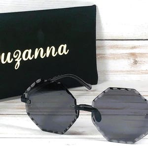 May include: Black sunglasses with a geometric octagon shape frame and dark gray lenses. The sunglasses are on a white surface next to a black case with the name "Suzanna" printed in white.