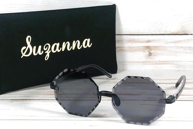 May include: Black sunglasses with a geometric octagon shape frame and dark gray lenses. The sunglasses are on a white surface next to a black case with the name "Suzanna" printed in white.