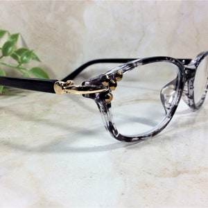Peut inclure: Lunettes dot&eacute;es d'une monture marbr&eacute;e gris et noir translucide avec une finition brillante. Les branches sont noir uni, reli&eacute;es &agrave; la face par des charni&egrave;res en m&eacute;tal dor&eacute;. Trois petits clous dor&eacute;s sont incrust&eacute;s sur chaque c&ocirc;t&eacute; de la monture.