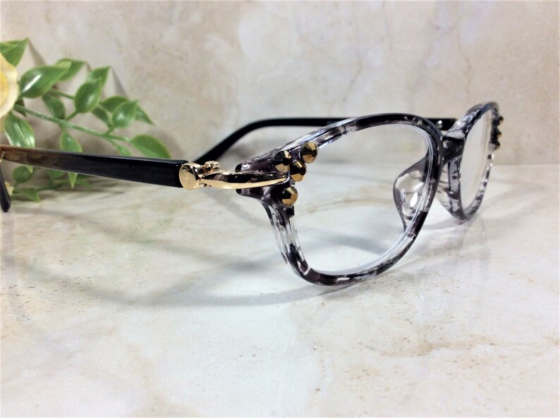 Peut inclure: Lunettes dot&eacute;es d'une monture marbr&eacute;e gris et noir translucide avec une finition brillante. Les branches sont noir uni, reli&eacute;es &agrave; la face par des charni&egrave;res en m&eacute;tal dor&eacute;. Trois petits clous dor&eacute;s sont incrust&eacute;s sur chaque c&ocirc;t&eacute; de la monture.