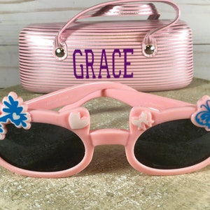 May include: Pink sunglasses with black lenses, adorned with blue flower and butterfly accents. A matching pink case with the name "GRACE" in purple lettering is in the background. The sunglasses are designed for children.