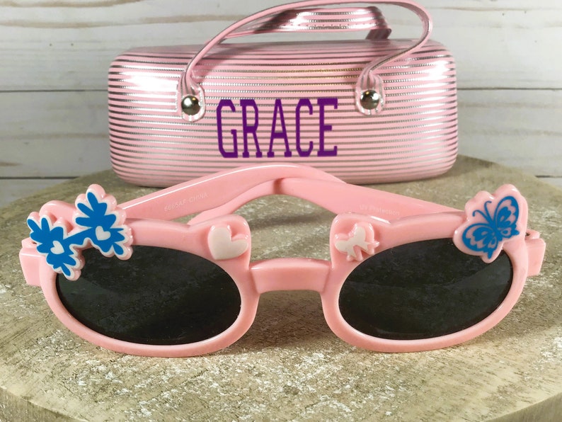 May include: Pink sunglasses with black lenses, adorned with blue flower and butterfly accents. A matching pink case with the name "GRACE" in purple lettering is in the background. The sunglasses are designed for children.