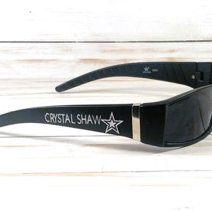 May include: Black sunglasses with a rectangular frame and curved temples. The text "CRYSTAL SHAW" is printed on the left temple, with a star graphic. A silver accent is on the frame.