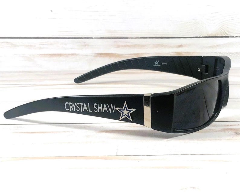 May include: Black sunglasses with a rectangular frame and curved temples. The text "CRYSTAL SHAW" is printed on the left temple, with a star graphic. A silver accent is on the frame.