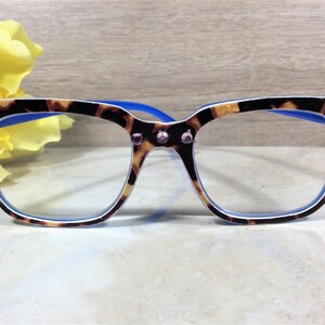 May include: Eyeglasses with a tortoiseshell pattern frame, blue arms, and clear lenses. The frame is adorned with pink rhinestones at the corners. A yellow flower is in the background. The glasses are on a white surface.