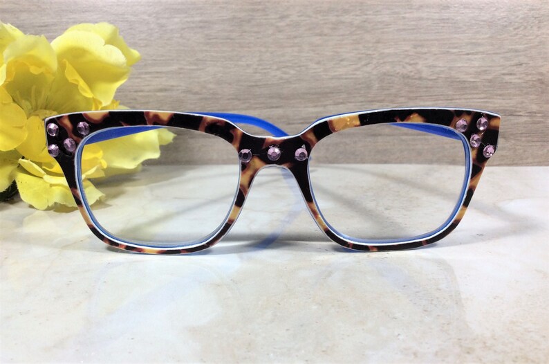 May include: Eyeglasses with a tortoiseshell pattern frame, blue arms, and clear lenses. The frame is adorned with pink rhinestones at the corners. A yellow flower is in the background. The glasses are on a white surface.