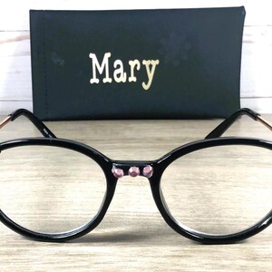 May include: Black-framed eyeglasses with clear lenses, a cat-eye design, and pink rhinestones. The frames have gold-coloured accents, and a black case with the name "Mary" in gold lettering.
