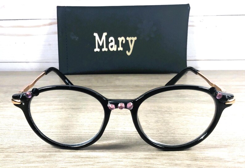May include: Black-framed eyeglasses with clear lenses, a cat-eye design, and pink rhinestones. The frames have gold-coloured accents, and a black case with the name "Mary" in gold lettering.