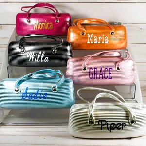 May include: A selection of striped eyeglass cases in shades of pink, orange, black, blue, and white. Each case has a handle and a name in a contrasting colour. The cases are designed for storing glasses and are personalised.