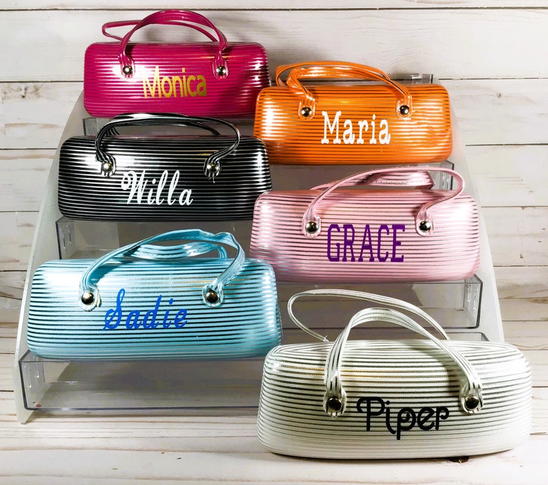 May include: A selection of striped eyeglass cases in shades of pink, orange, black, blue, and white. Each case has a handle and a name in a contrasting colour. The cases are designed for storing glasses and are personalised.