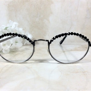 May include: Round eyeglasses with a silver frame and clear lenses. The top of the frames are embellished with a row of small, black, faceted beads. The glasses are set against a soft, blurred background with white flowers.