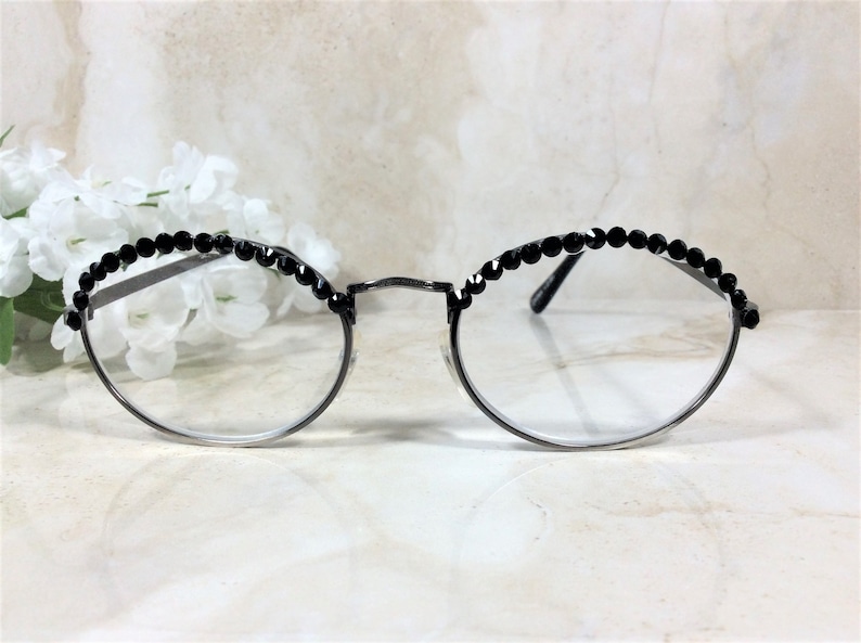 May include: Round eyeglasses with a silver frame and clear lenses. The top of the frames are embellished with a row of small, black, faceted beads. The glasses are set against a soft, blurred background with white flowers.