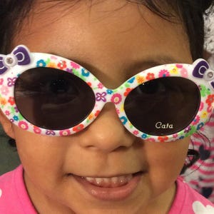 May include: A young girl wearing white sunglasses with a floral and colorful design. The sunglasses have a purple bow on each side and the text "Cata" is printed on the right lens.